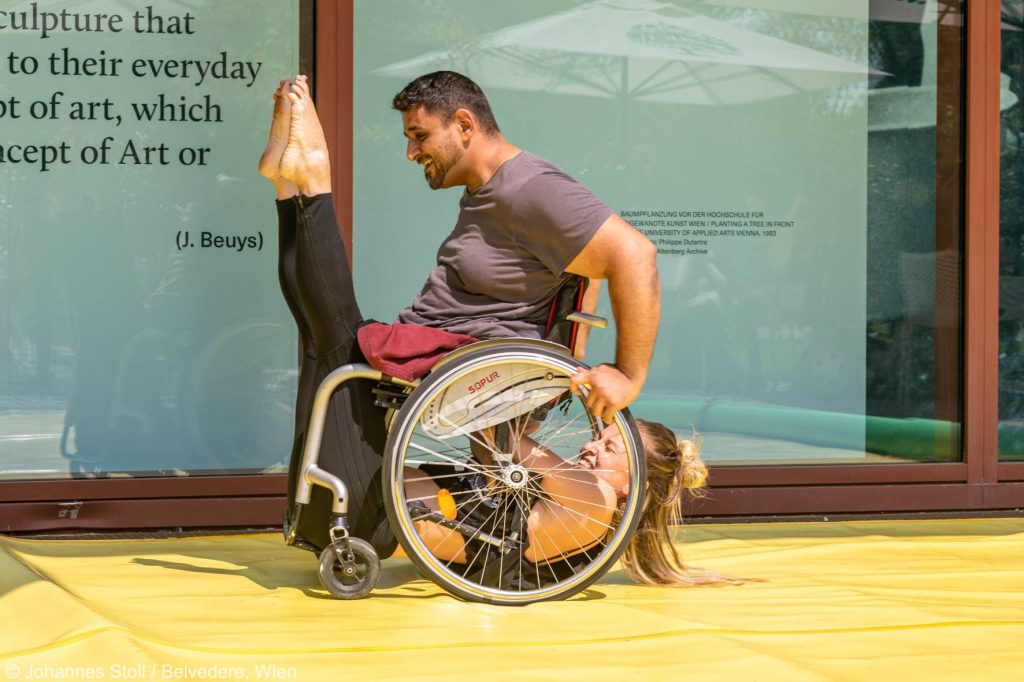Choreografie: Sie hängt unter dem Rollstuhl, er fährt und hat ihre Füße vor dem Gesicht.