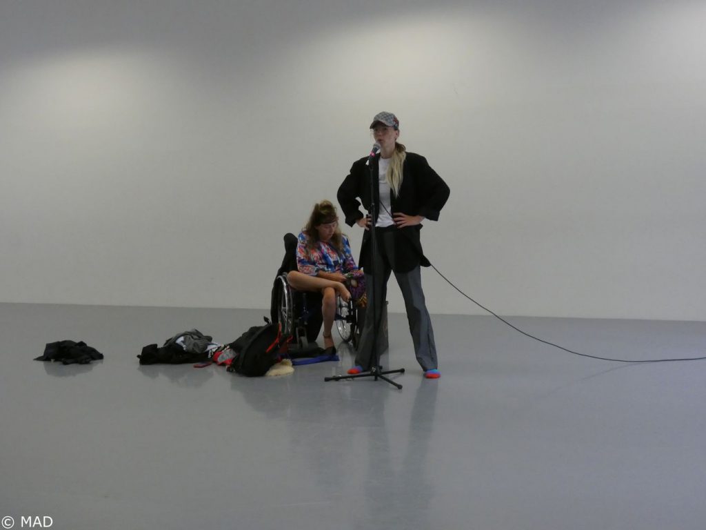 Eine der zwei Künstler*innen betrachtet im Rollstuhl sitzend ein Kleidungsstück. Die andere der Künstler*innen steht selbstbewusst davor am Mikrophon.