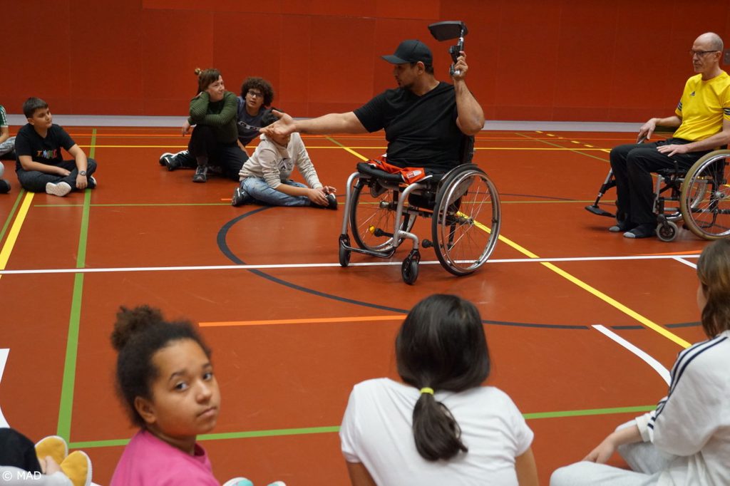 Adil Embaby zeigt die einzelnen Teile eines Rollstuhls, die Schüler*innen sitzen im Kreis und hören gespannt zu.