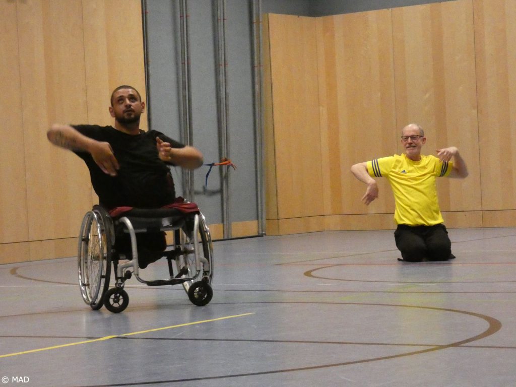 Zwei Künstler tanzen im Turnsaal. Adil Embaby, einer der beiden Künstler, nutzt einen Rollstuhl. Frans Poelstra im gelben T-Shirt kniet neben ihm. Sie begegnen sich auf gleiche Augenhöhe.