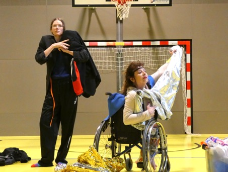 Zwei Künstler*innen kostümieren sich auf der Bühne. Eine der beiden Personen steht (links), die andere sitzt in ihrem Rollstuhl (rechts).