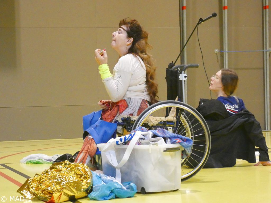 Im Vordergrund trägt eine Künstlerin im Rollstuhl Lippenstift auf. Im Hintergrund posiert ein*e zweite KÜnstler*in liegend.