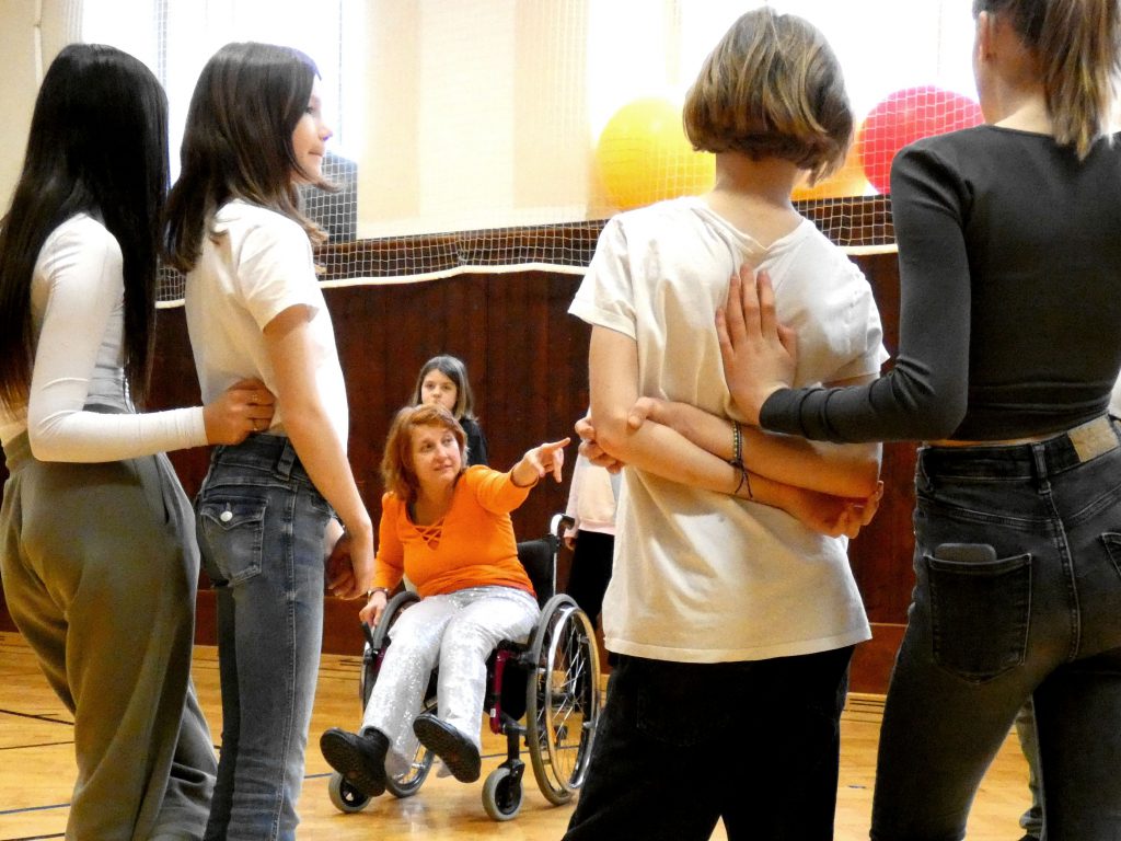 Eine Tänzerin im Rollstuhl fordert eine Schülerin zu etwas auf. Vier Jugendliche sehen konzentriert zu.