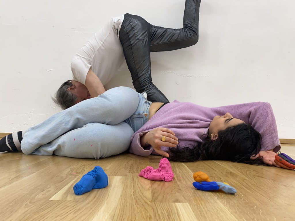Zwei Künstler*innen tanzen an Boden hinter kleinen bunten Skulpturen aus Plastilin. Sie sehen dabei selbst aus wie Kunstwerke.